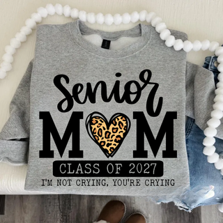 Flat lay of a heather grey crewneck sweatshirt with a black Senior MOM design, a leopard print heart, a Class of 2027 banner, and the tagline I'm not crying, you're crying