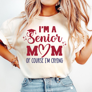 Close-up of a woman in an ivory tee with a red I'm a Senior MOM design featuring a graduation cap, heart, sparkles, and navy Of course I'm crying lettering