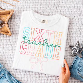 White Comfort Colors sixth grade teacher bow graphic tee with retro varsity lettering flat lay
