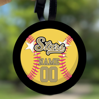  Black round bag tag held up outdoors featuring yellow softball with pink stitching gold script Stars logo white stars and personalized gold name and number DTF print with black hanging strap on blurred green background