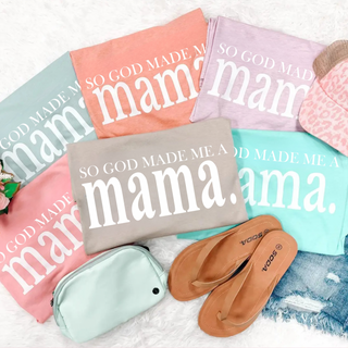 So God Made Me a Mama graphic tees in dusty blue, sunset, lilac, pink, tan, and mint laid flat with sandals and accessories