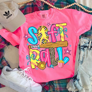 Comfort Colors neon pink short sleeve tee laid flat with colorful doodle softball graphic featuring player bat glove bows leopard print and checkered letters styled with jeans and white sneakers