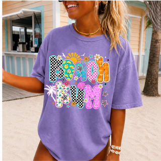 violet Comfort Colors tee with colorful beach bum checkered patterned letter design, worn by smiling woman at the beach
