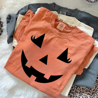 Stacked fall tops featuring the Sweet & Spooky Jack-O-Lash Graphic Top in a warm orange tone with bold black jack-o-lantern face and lash detail, layered with neutral and charcoal garments for Halloween styling inspiration.