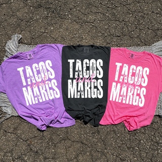 Three tees laid flat in grape, black, and pink each showing TACOS and MARGS distressed block letter graphic with pink and script accent
