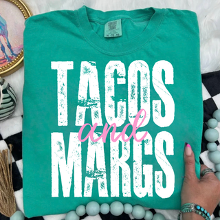 Island green tee folded flat showing TACOS and MARGS in large distressed block letters with pink and script accent
