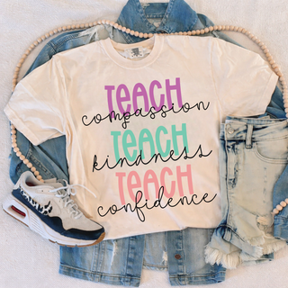 White tee flat lay with TEACH compassion TEACH kindness TEACH confidence stacked colorful script design
