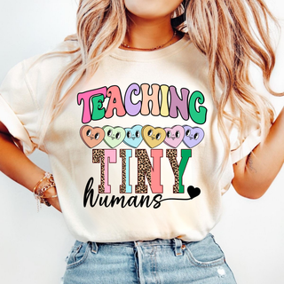 Woman wearing an ivory tee with TEACHING TINY humans colorful groovy leopard print heart graphic on the front

