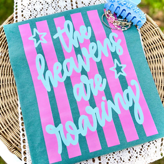 Seafoam Comfort Colors tee with bold pink stripe background and the words the heavens are roaring in light blue script with stars laid on a wicker basket outdoors