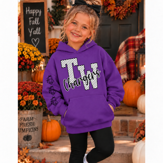 Girl in a purple hoodie with white polka dot TW letters outlined in black and black Chargers script on the front, with three black charger horse head graphics down the sleeve, standing on a fall porch with pumpkins