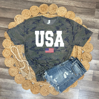Green camouflage USA shirt styled flat lay with bold white varsity USA lettering and small American flag graphic underneath, paired with distressed denim shorts on a round woven jute rug with wood bead tassel decor on a wood floor background