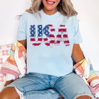 Woman wearing a chambray tee with a bold USA faux patch graphic in american flag stars and stripes letters on the front
