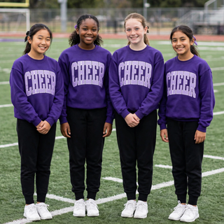 Four youth cheerleaders wearing matching purple crewneck sweatshirts with light lavender arched Cheer graphic standing on football field