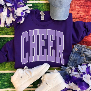 Flat lay of dark purple Cheer sweatshirt with lavender varsity arched Cheer text styled with purple white pom poms denim cap and white sneakers on turf