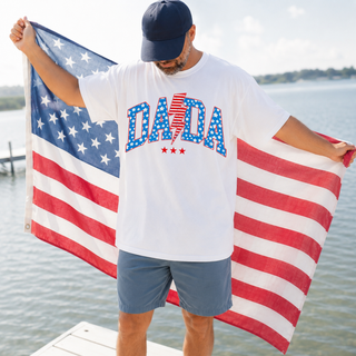 man wearing white tee with dada stars and stripes lightning bolt design, holding American flag on a dock by the water
