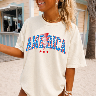 ivory Comfort Colors tee with america stars stripes lightning bolt design, worn by smiling woman at a beach boardwalk
