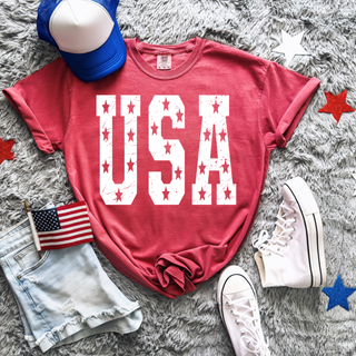 Crimson Comfort Colors adult tee with distressed white USA text and stars flat lay with blue trucker hat, denim shorts, American flag, white high top sneakers, and red white blue star decor
