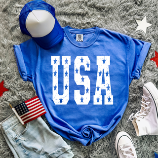 Flo Blue Comfort Colors adult tee with distressed white USA text and stars flat lay with blue trucker hat, denim shorts, American flag, white high top sneakers, and red white blue star decor