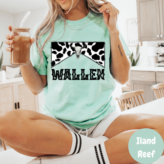 woman wearing island reef garment dyed cotton tee with black and white cow print bow, longhorn skull, and bold wallen text holding iced coffee