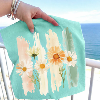 Hand holding a Chalky Mint Comfort Colors top with a watercolor daisy and brushstroke graphic in peach cream and sage with ocean in background