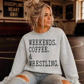 Woman in a light grey crewneck sweatshirt with stacked black typewriter-style text reading Weekends. Coffee. & Wrestling., seated in a wrestling room