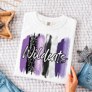 Folded white Comfort Colors tee on a gray knit blanket showing purple, black, and silver glitter leopard brushstrokes with white script Wildcats lettering outlined in black