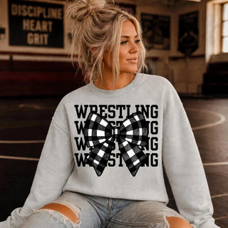 Woman in a heather grey crewneck sweatshirt with stacked black Wrestling lettering and a black and white buffalo plaid bow, seated in a wrestling room