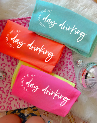 Folded tees in mint, coral, neon pink, and yellow each showing YOU HAD ME AT day drinking diagonal script graphic
